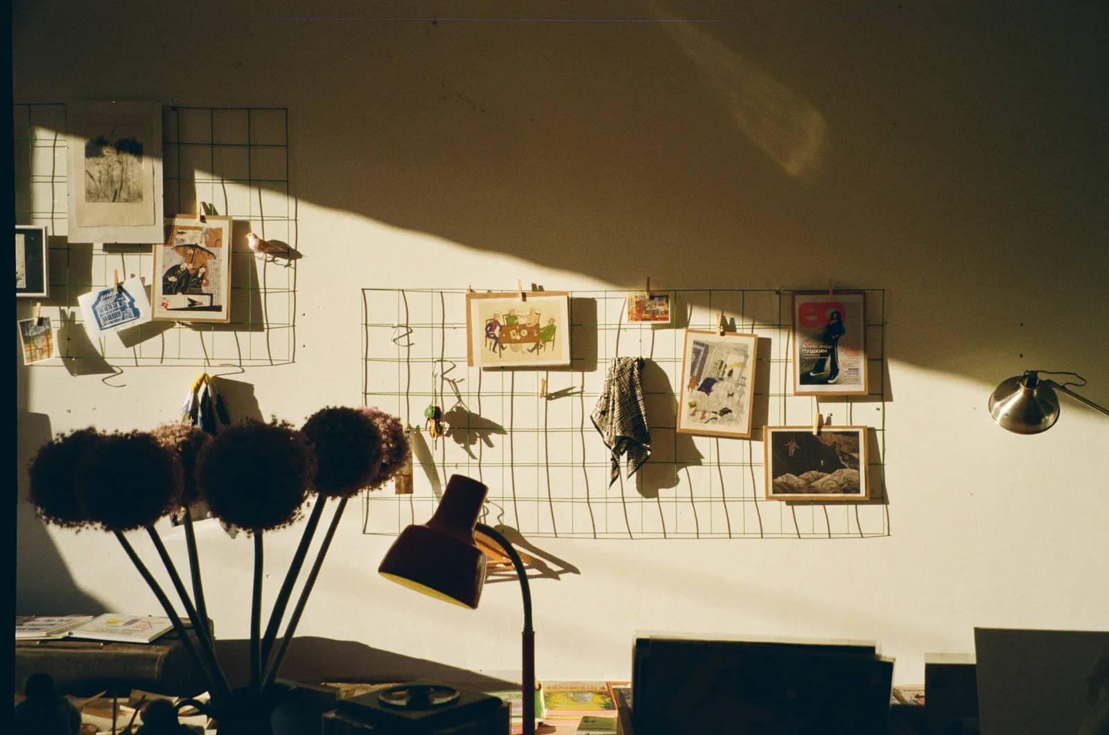 Small art prints and postcards clipped to wire grid panels on a sunlit studio wall above a desk with dried flowers and a red lamp