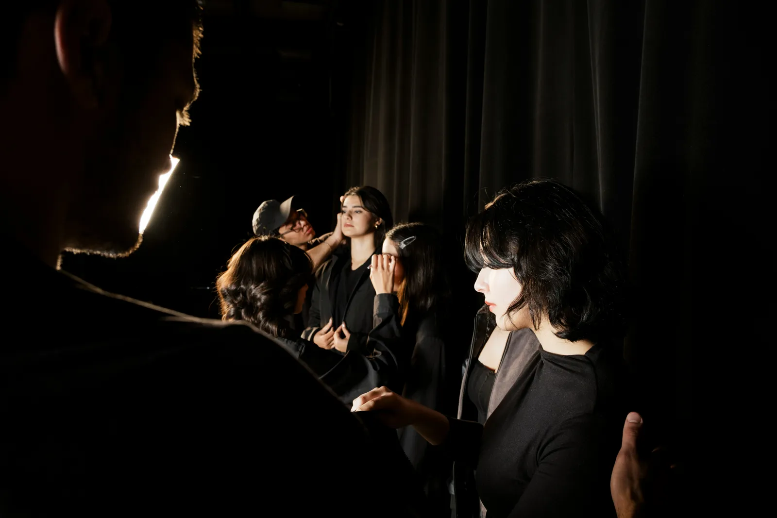 Makeup artists working on models in black outfits backstage behind dark curtains, lit by a single bright light