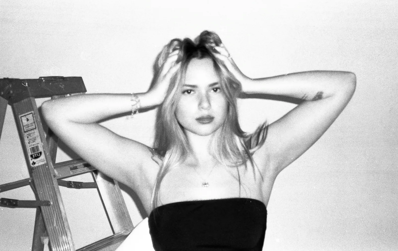 Black and white film photo of a young woman in a strapless top with her hands in her hair next to a wooden step ladder