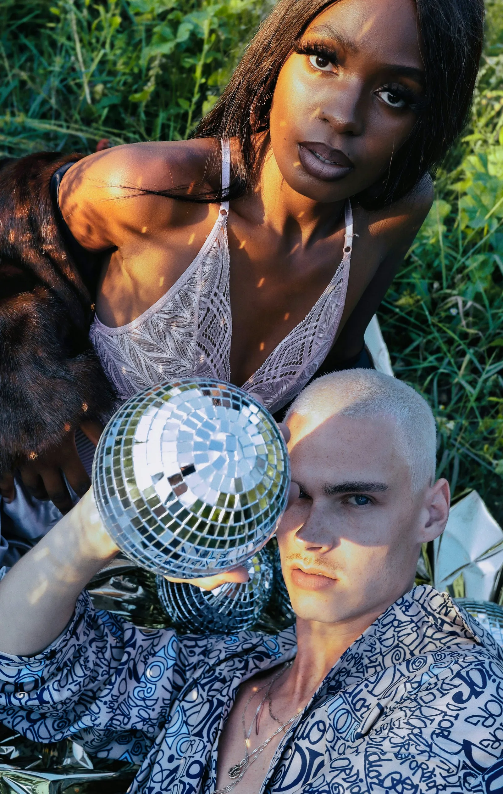 Woman in a lace top and a man with bleached hair in a patterned shirt holding a disco ball while sitting in tall grass