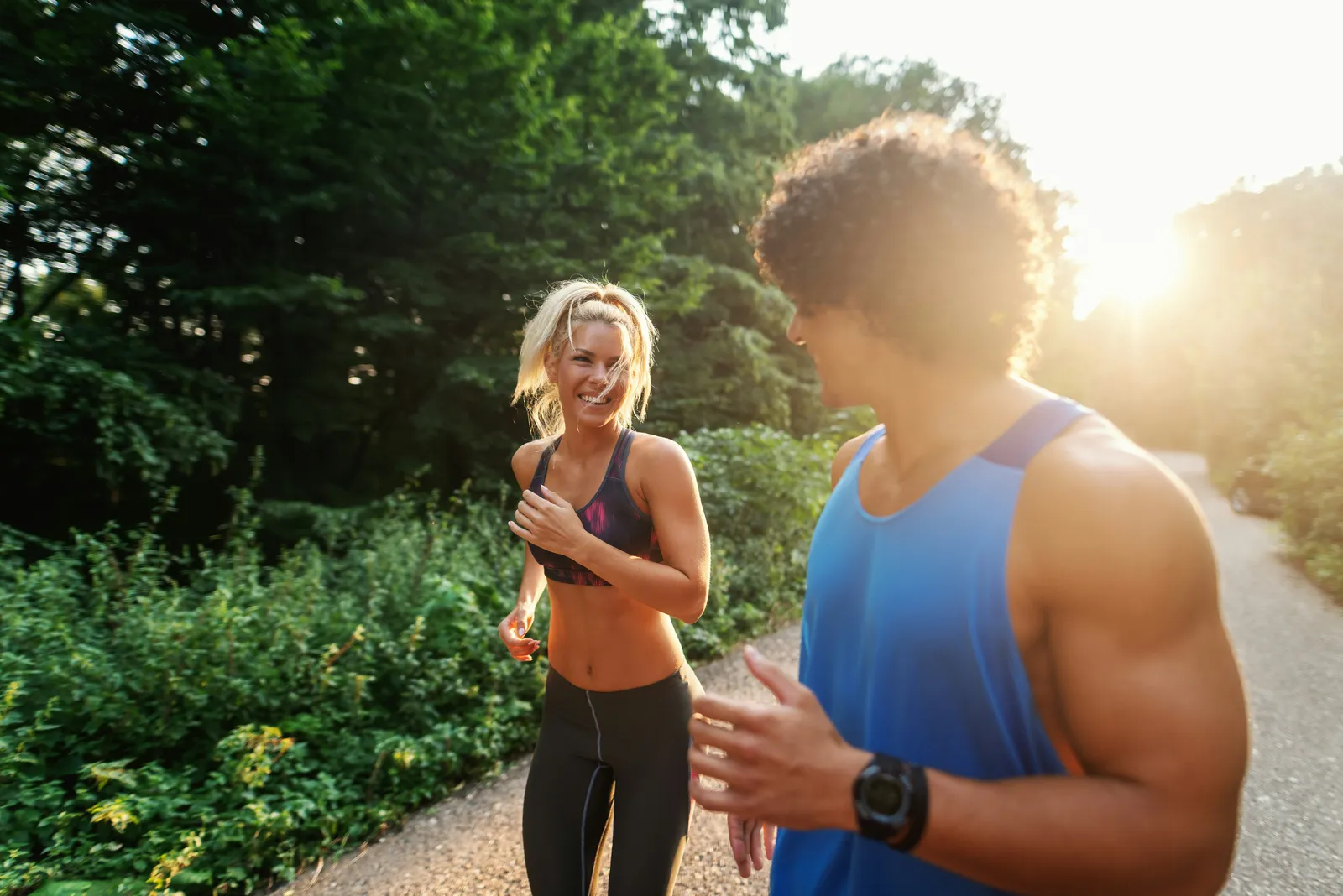 Smiling woman in a sports bra and a man in a blue tank top jogging along a tree-lined path in warm sunlight