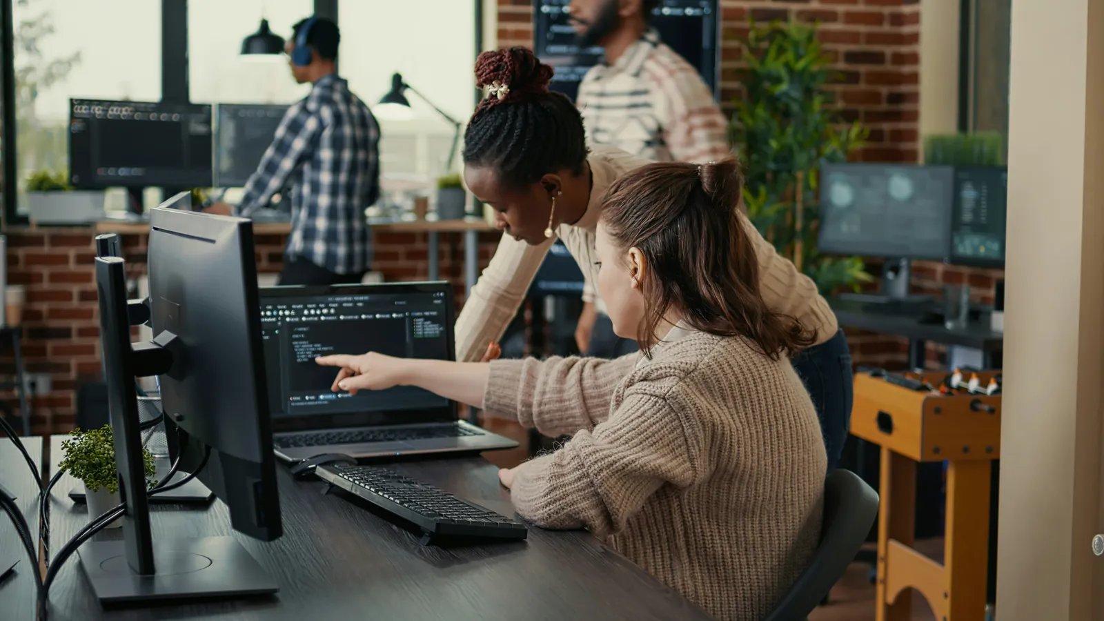 Two developers at a desk pointing at code on a laptop screen in an office with brick walls and more programmers behind them