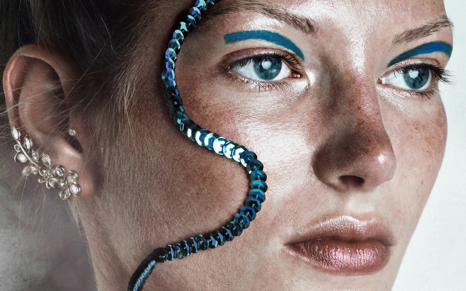 Close-up of a freckled face with blue eyeliner, a strip of blue sequins curving across the cheek and a crystal ear cuff