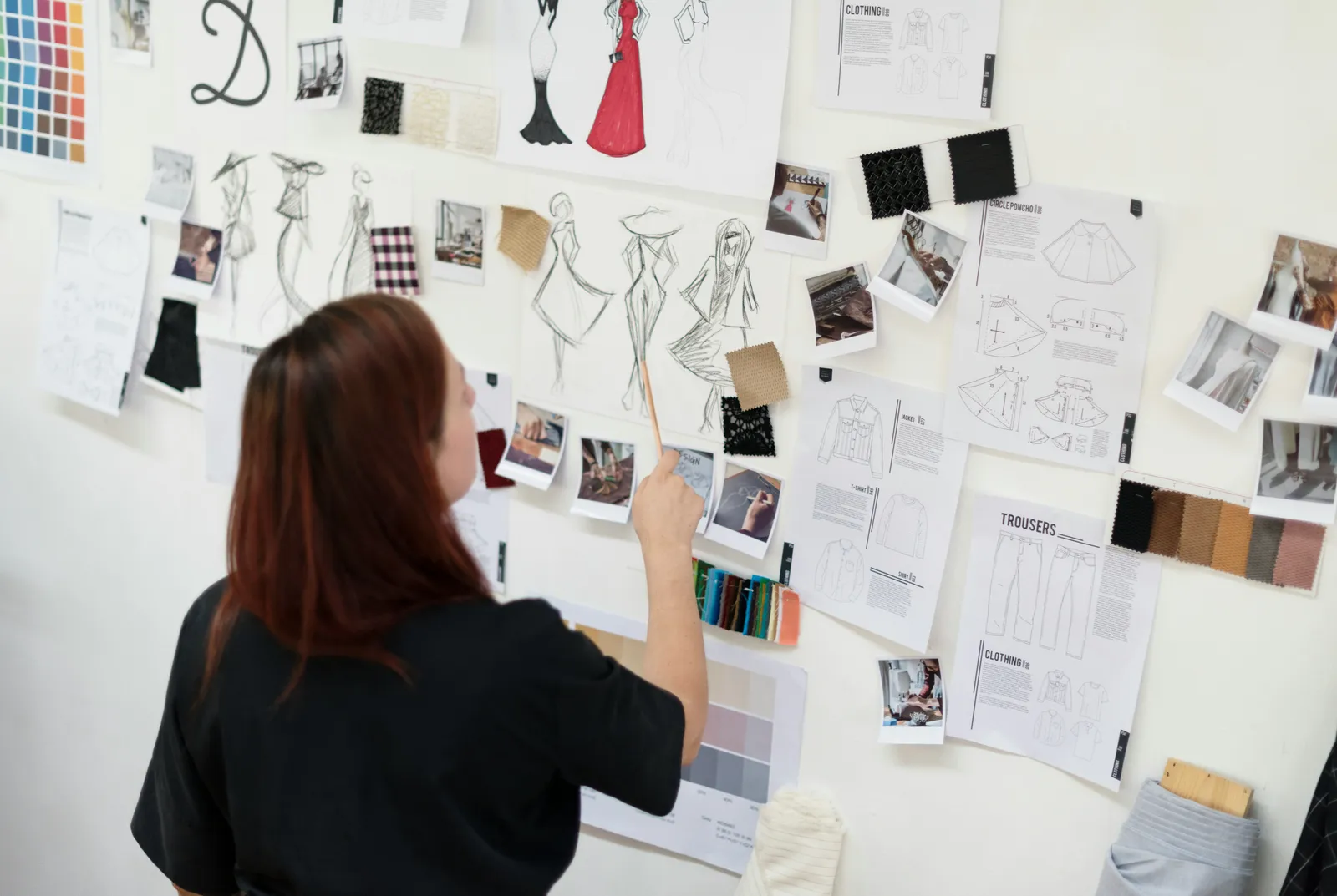 Fashion designer with red hair pointing at clothing sketches, fabric swatches and garment spec sheets pinned to a studio wall