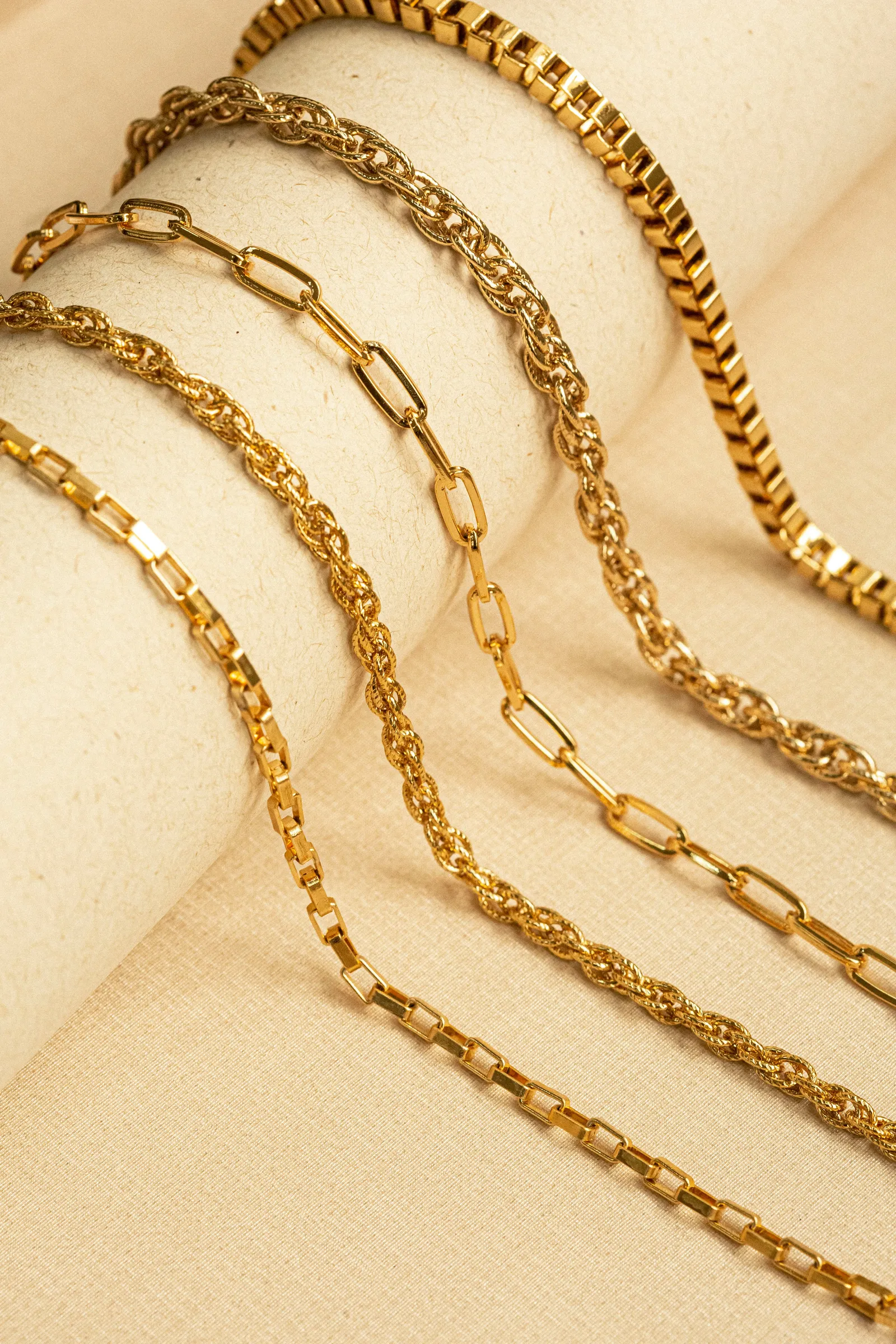 Five different gold chain necklaces draped side by side over beige paper rolls