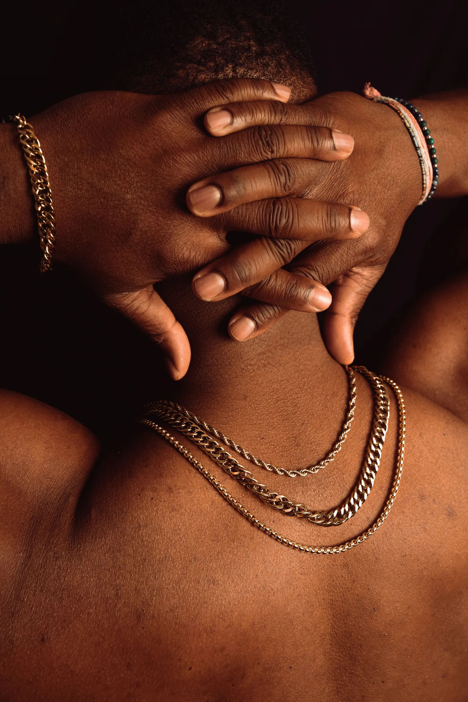 Back view of a man with hands clasped behind his neck wearing three layered gold chain necklaces and a gold bracelet