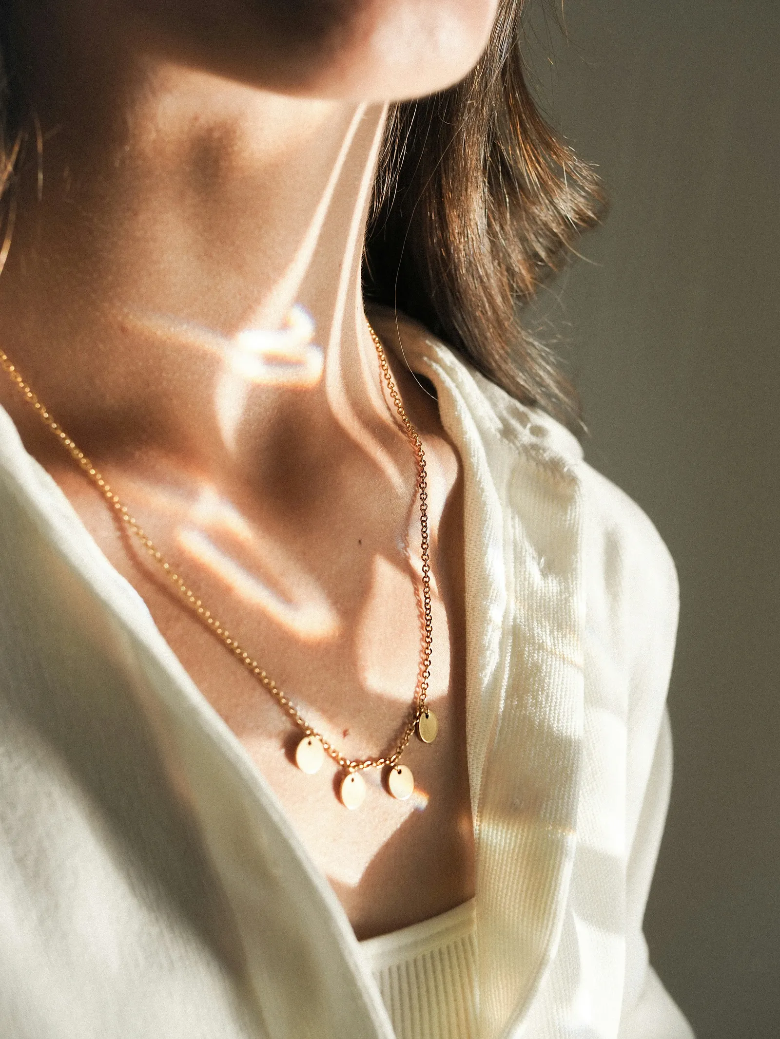 Close-up of a delicate gold chain necklace with small disc pendants on a woman's neck, lit by sunlight through a white blouse