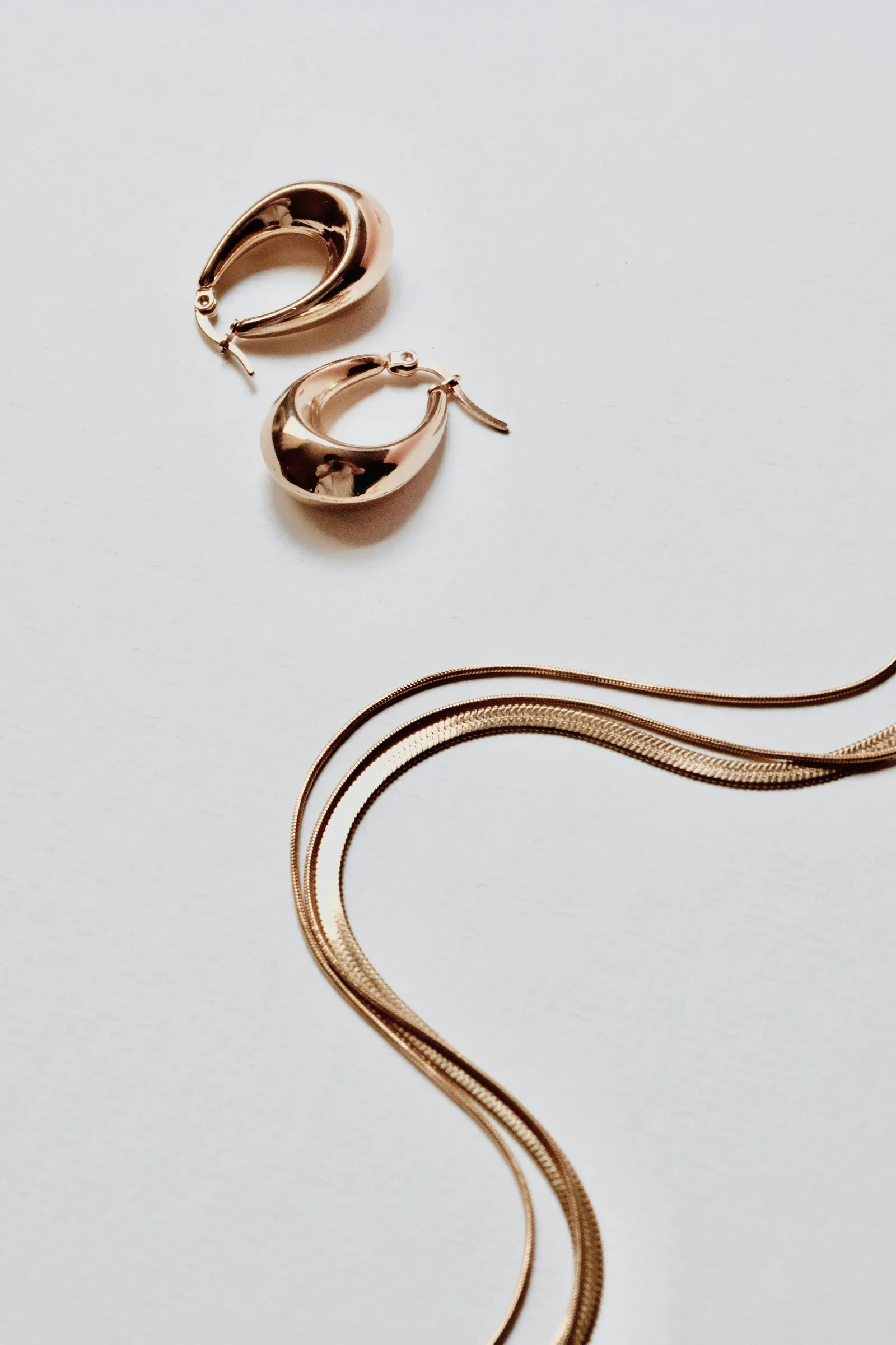 Pair of chunky gold hoop earrings and two gold herringbone chains on a white surface