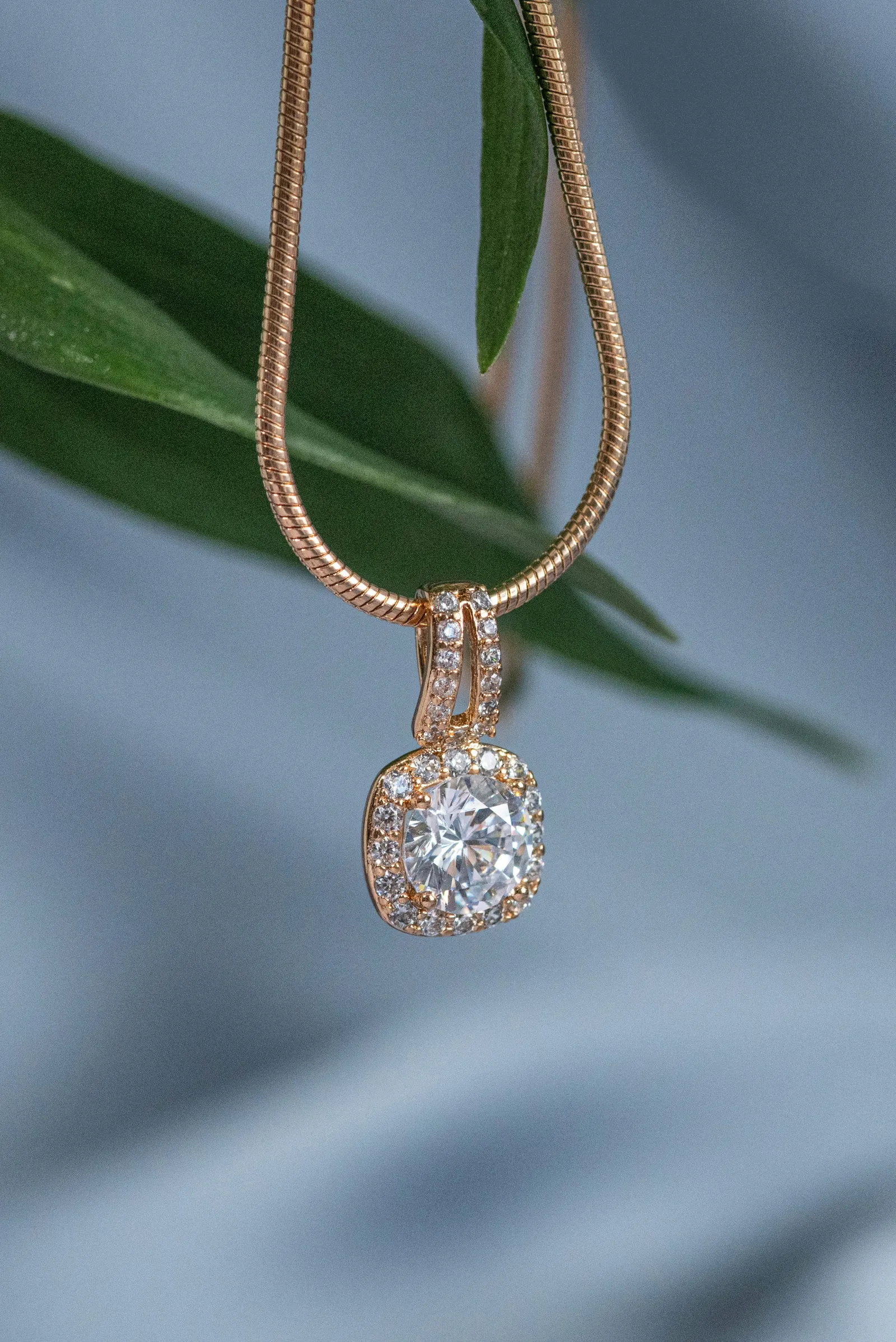 Gold chain necklace with a square crystal halo pendant hanging in front of a green leaf and soft blue fabric