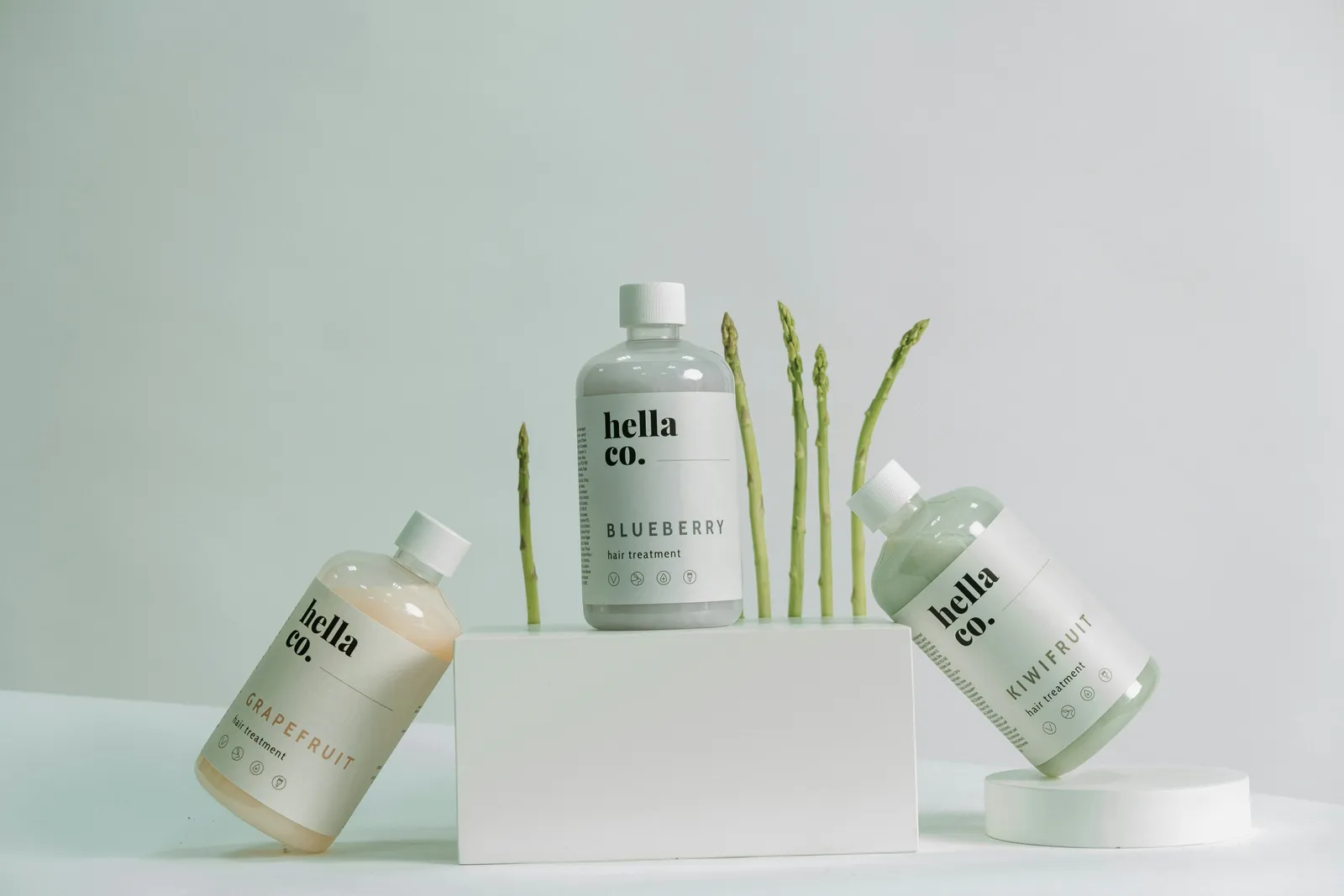 Three pastel hair treatment bottles with white labels arranged on white podiums with asparagus stems against a pale green background