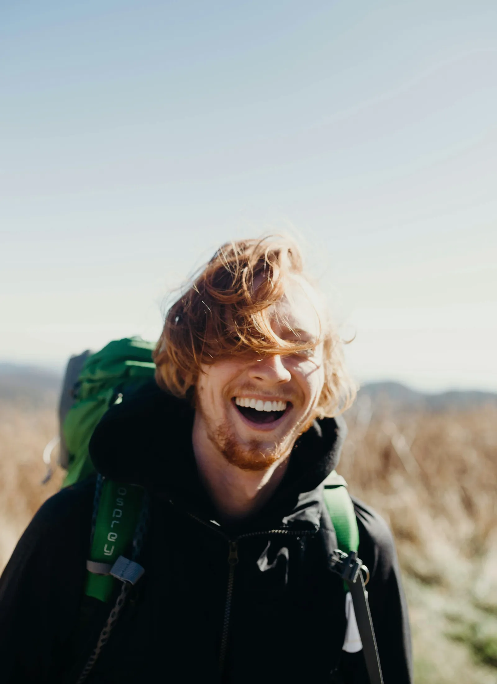Laughing red-haired man in a black jacket with a green hiking backpack on a windy mountain ridge