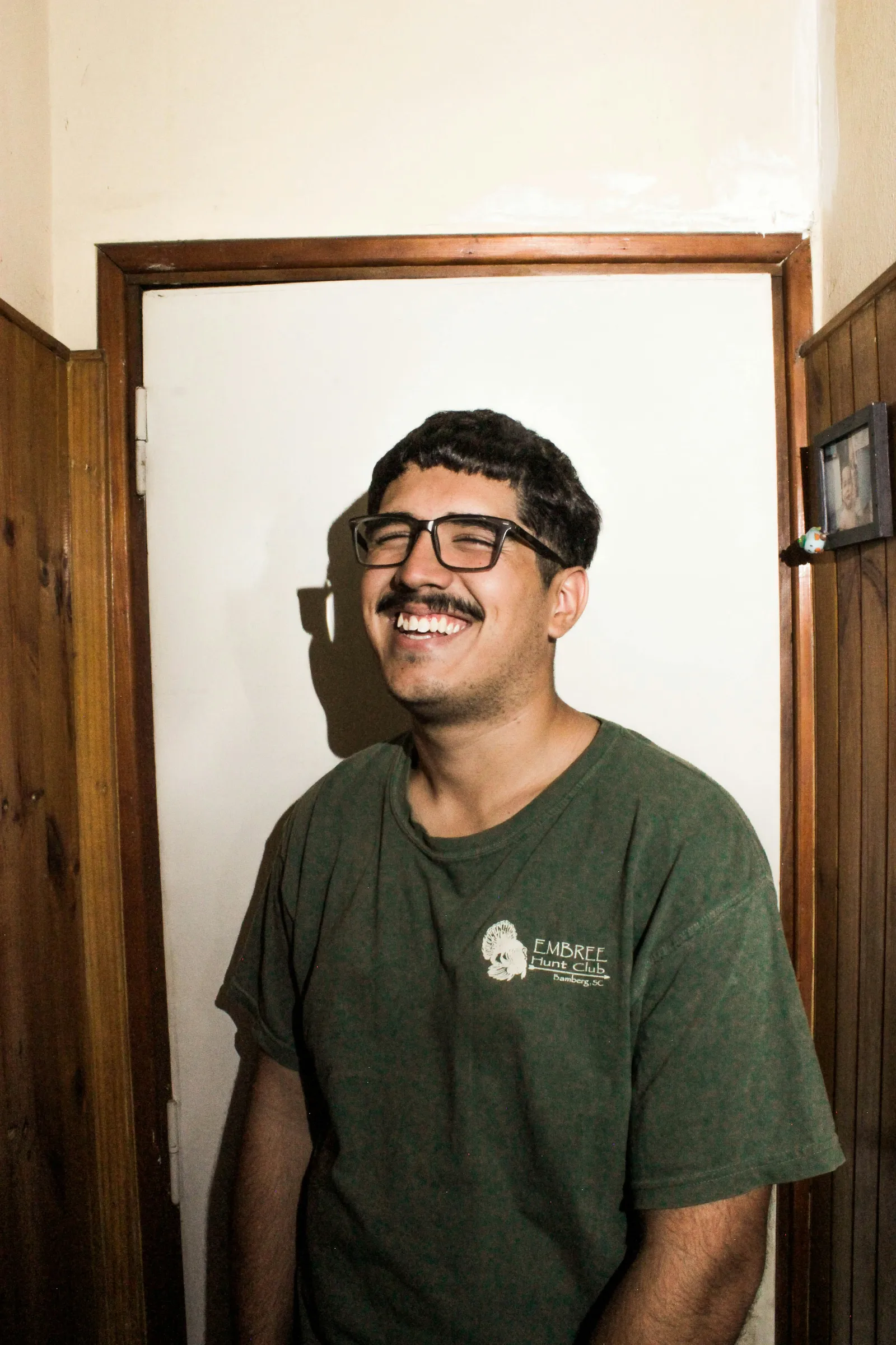Man with glasses and a moustache laughing in a green printed t-shirt in front of a white door, lit by camera flash