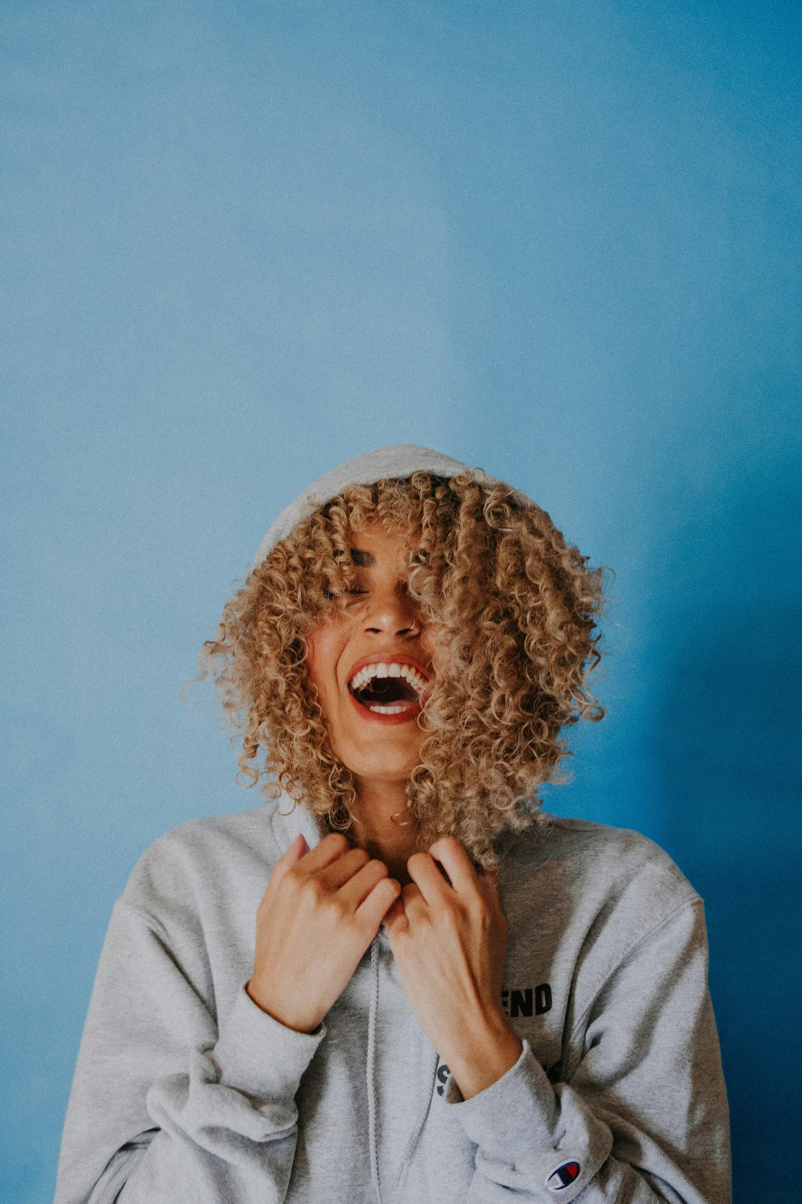 Laughing woman with blonde curly hair wearing a grey hoodie with the hood up against a blue wall