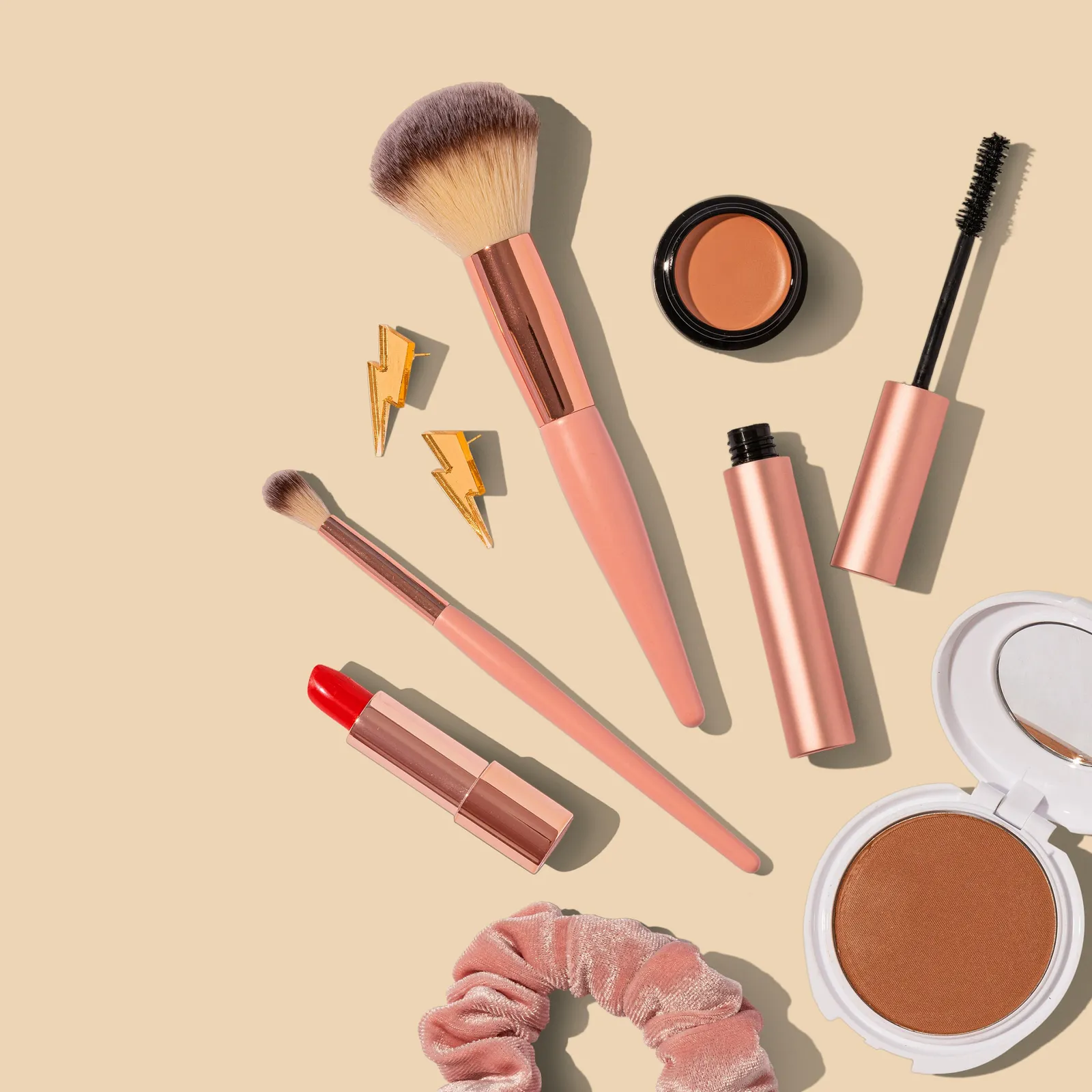 Flatlay of makeup brushes, red lipstick, mascara, a bronzer compact, a velvet scrunchie and lightning bolt earrings on a beige background