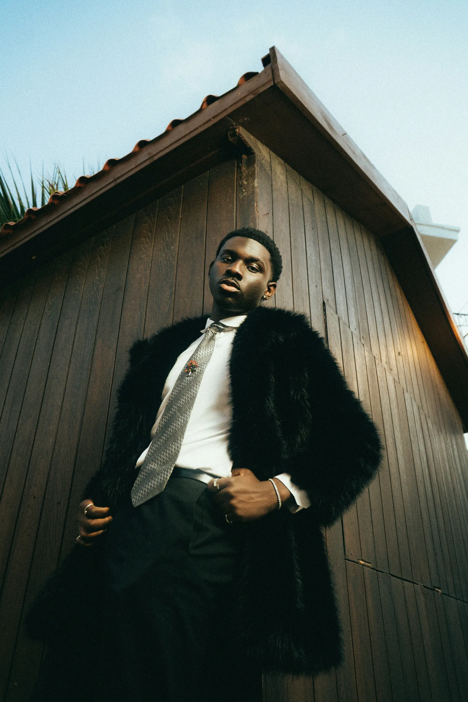 Low-angle portrait of a man in a black fur coat, white shirt and patterned tie leaning against a wooden cabin wall