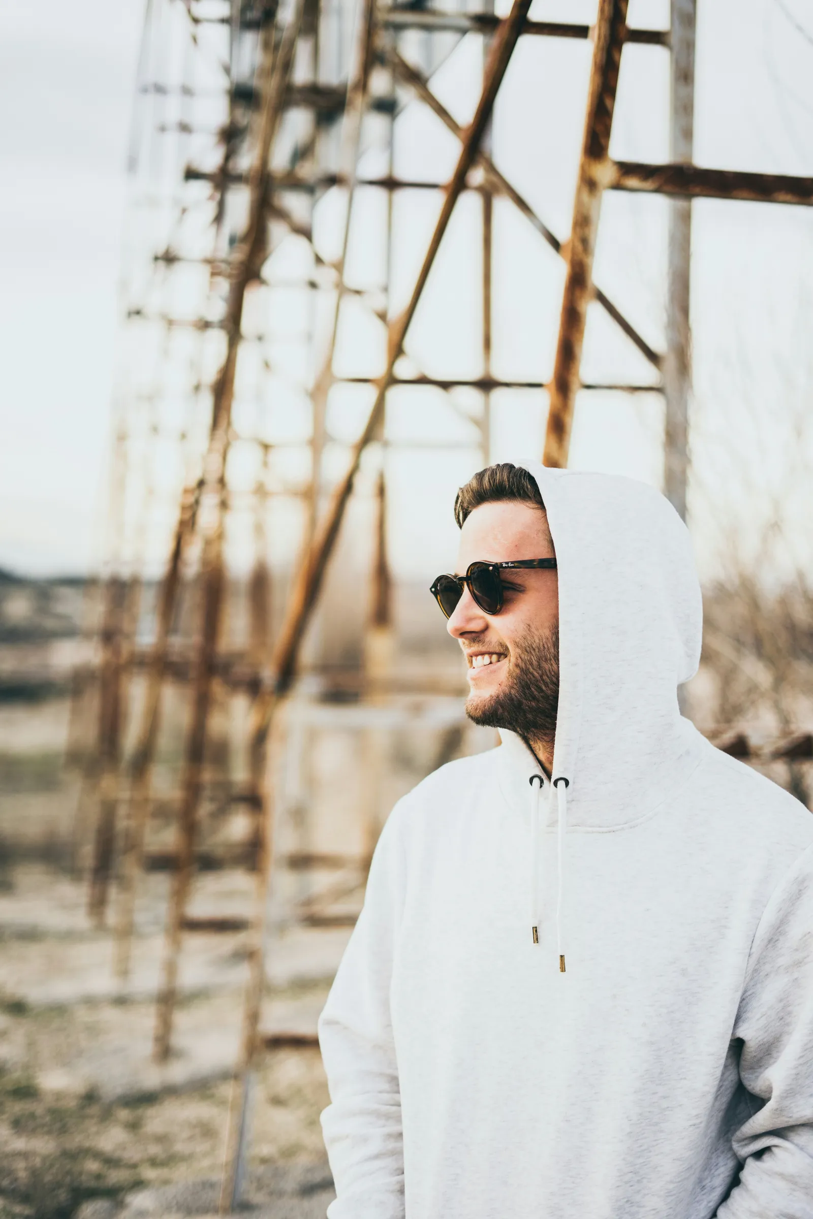 Smiling bearded man wearing a plain light grey hoodie with the hood up and dark sunglasses in front of a rusty metal tower