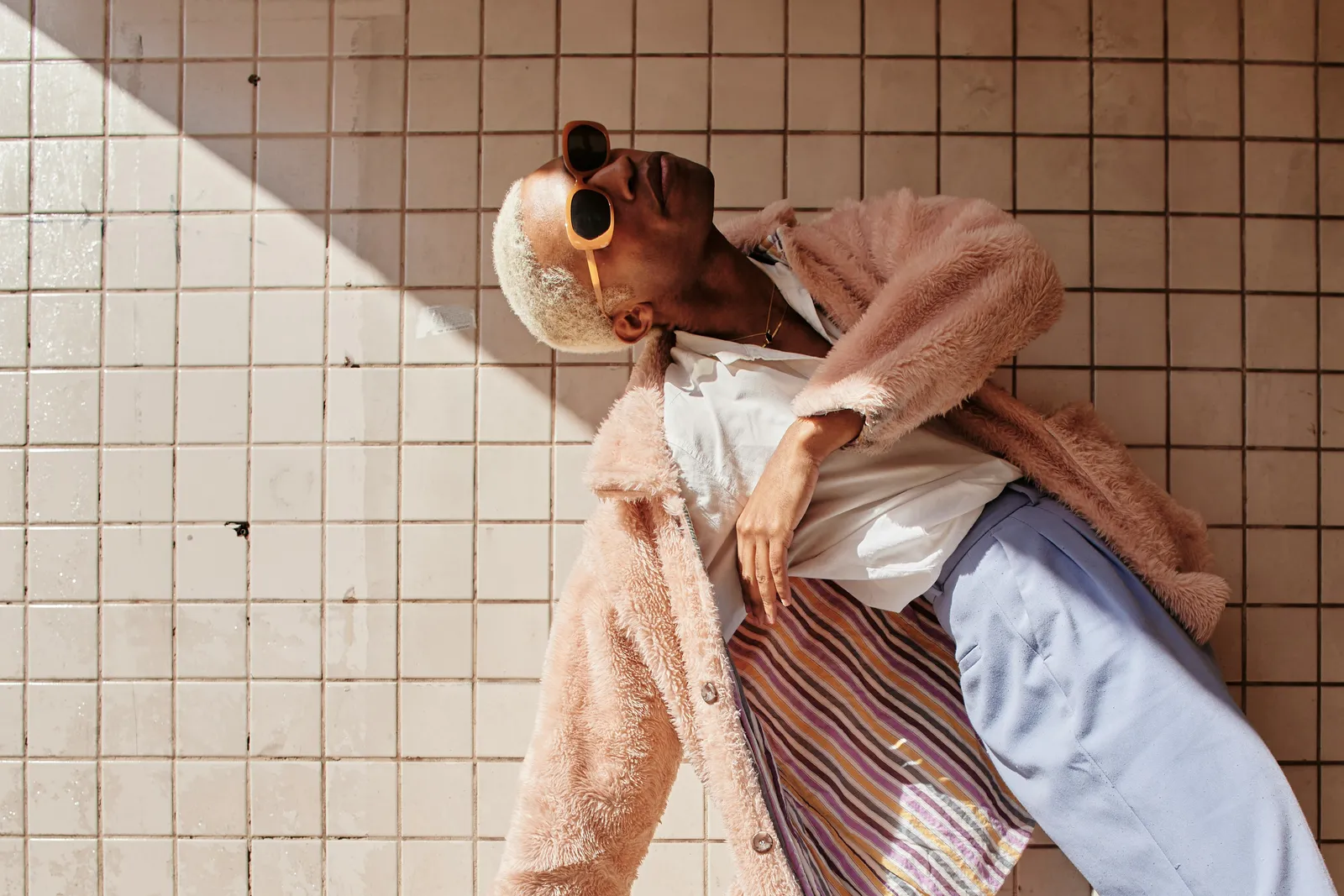 Man with bleached hair leaning back against a white tiled wall in a pink fluffy coat, orange sunglasses and striped trousers