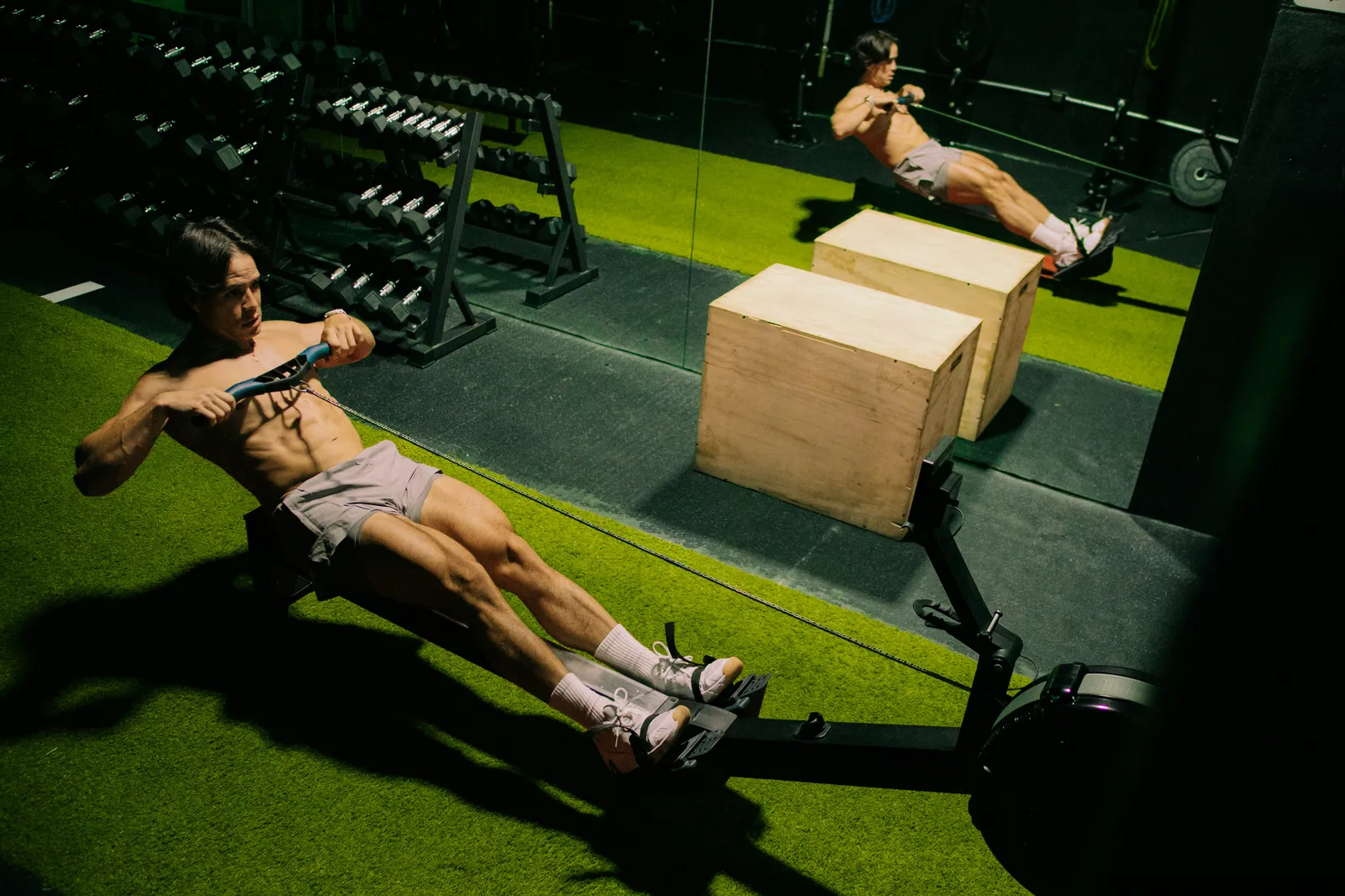 Shirtless man using a rowing machine on green turf in a gym with dumbbell racks and wooden plyo boxes
