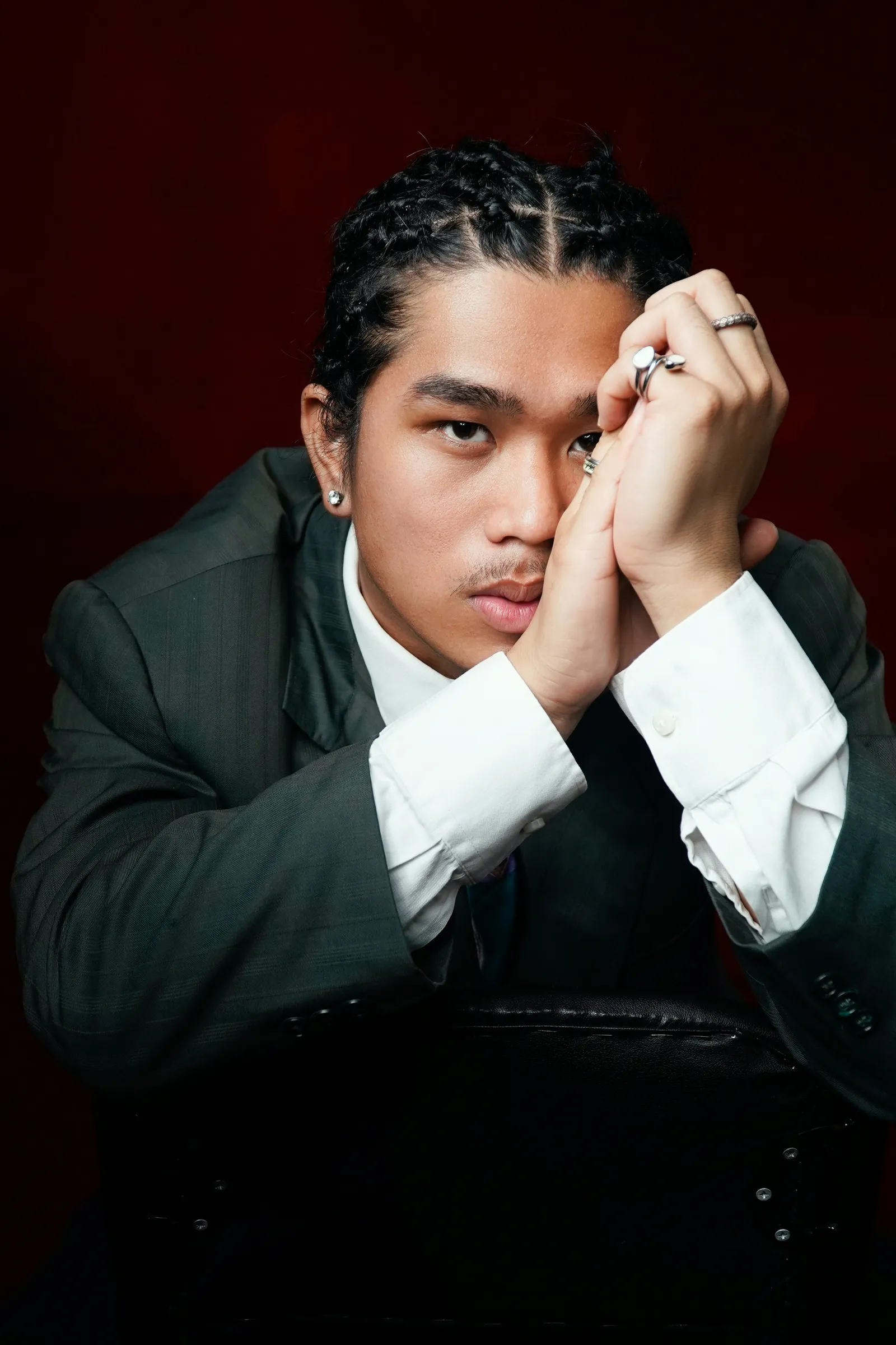 Man with braided hair in a dark suit resting his chin on clasped hands wearing silver rings against a deep red background