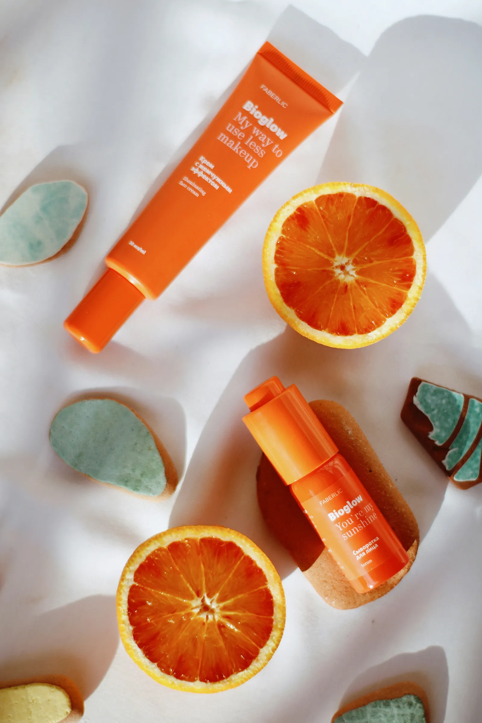 Flatlay of an orange skincare tube and serum bottle with halved oranges and pastel stones on white fabric