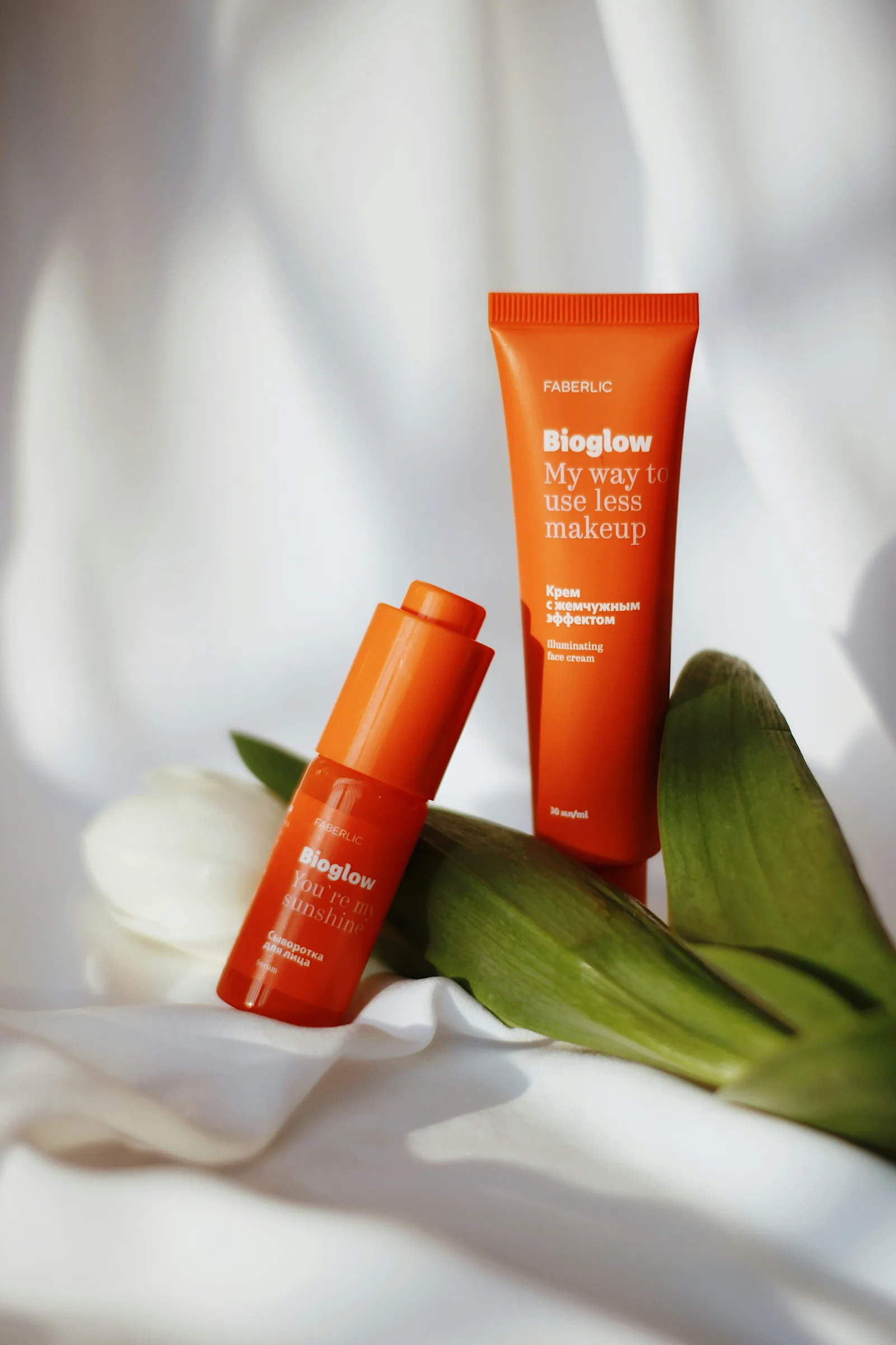 Orange skincare tube and small serum bottle standing on white fabric next to a white tulip