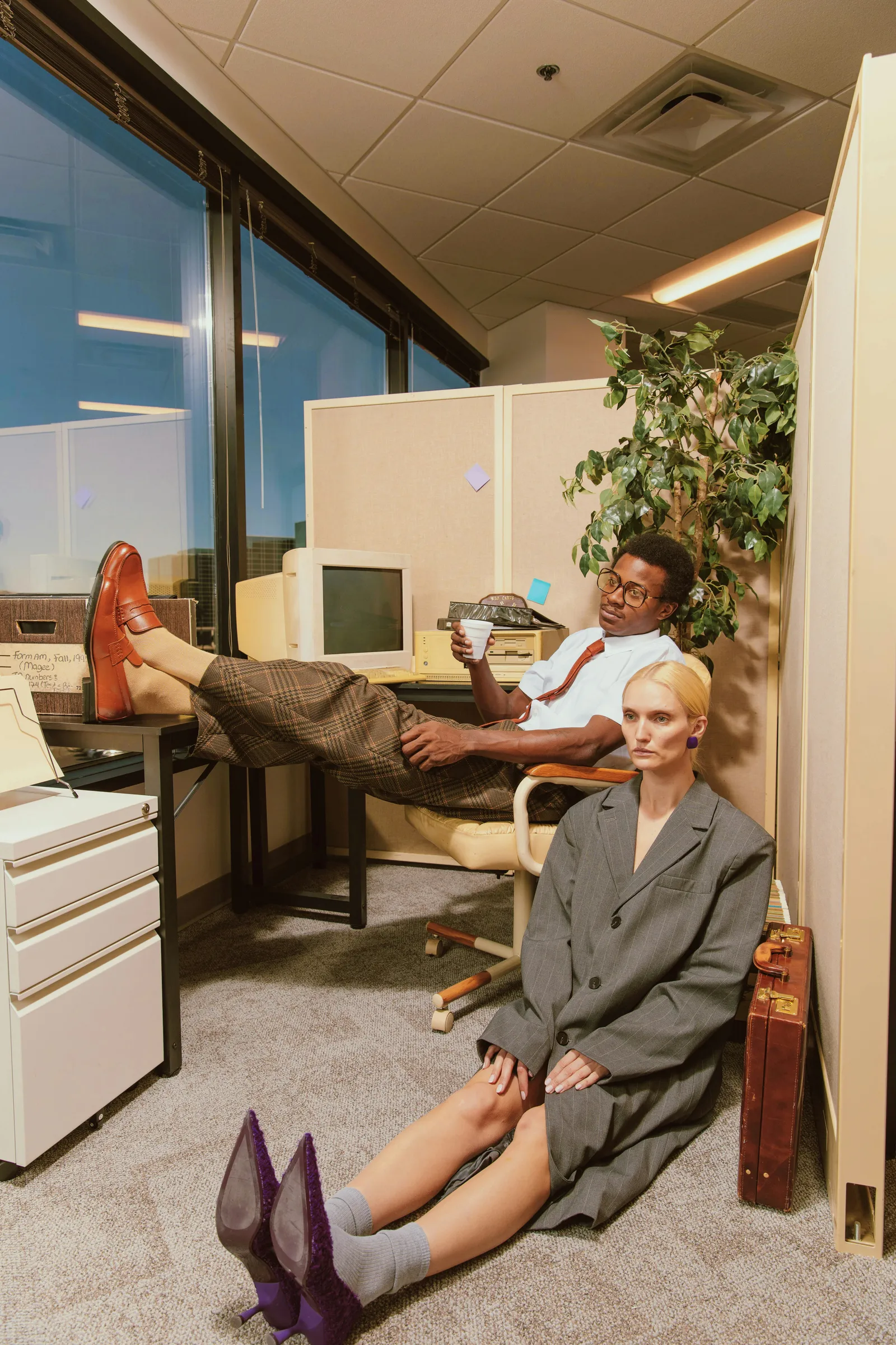 Fashion editorial in a retro office with a man resting his feet on a desk beside an old computer and a woman in an oversized grey blazer sitting on the carpet