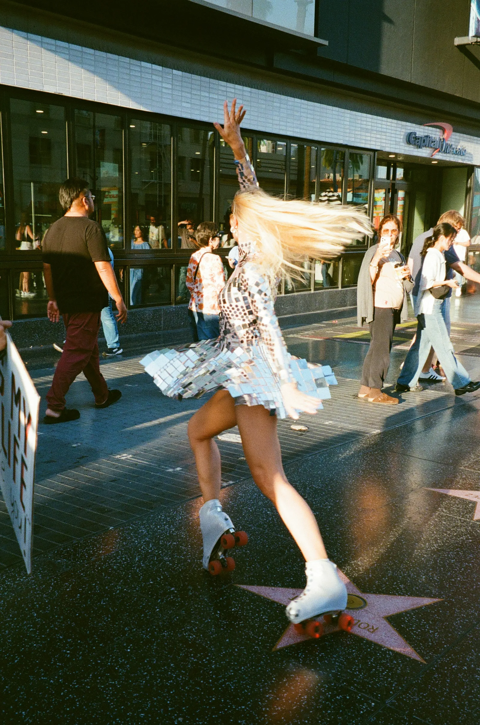 Woman in a silver sequin dress and white roller skates spinning with one arm raised on a busy city sidewalk