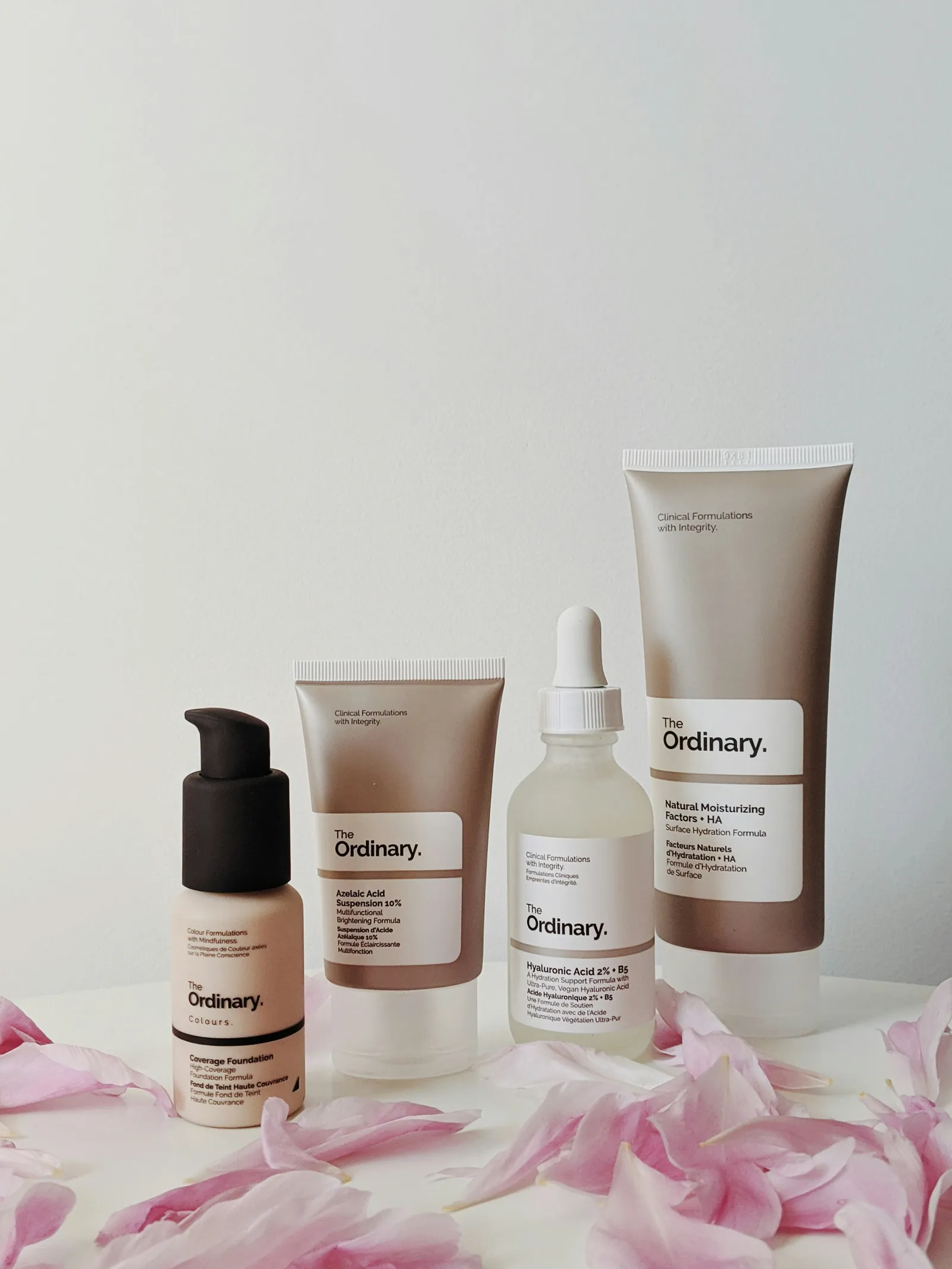 Four skincare products with white labels, including a dropper bottle and squeeze tubes, lined up on a white table scattered with pink petals