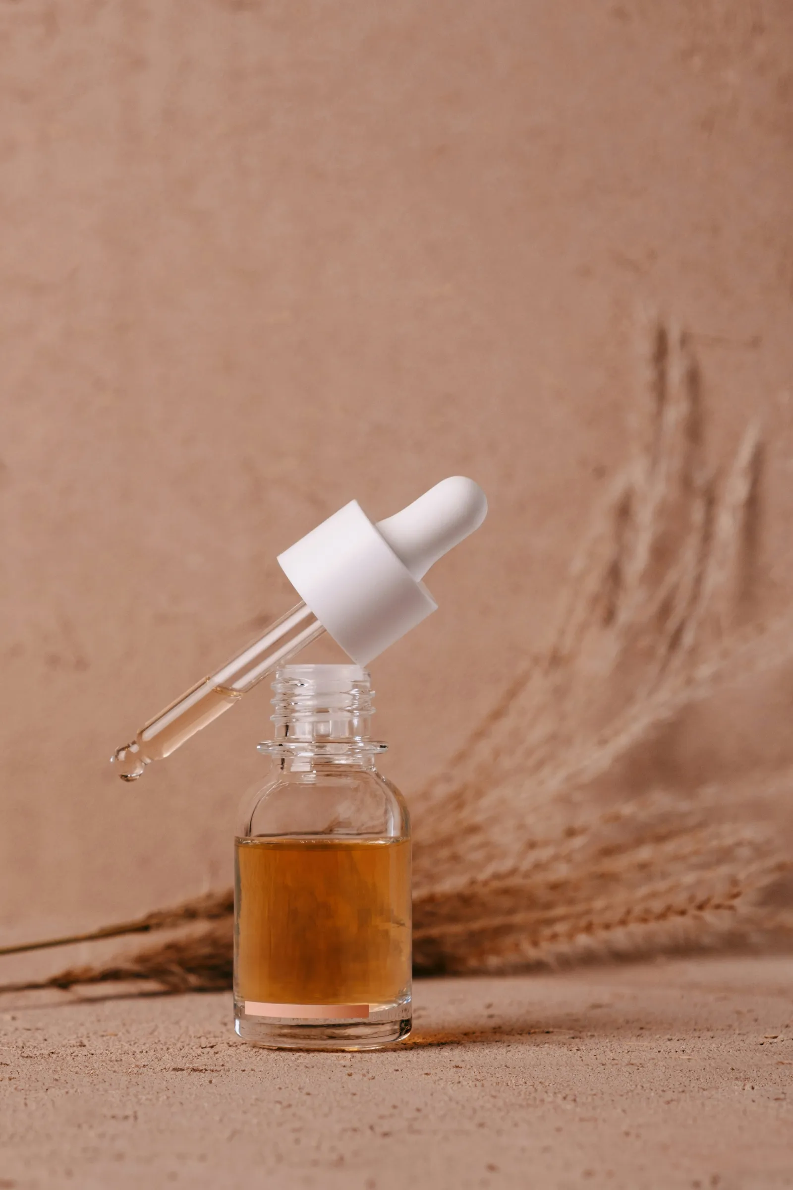 Glass dropper bottle of amber serum with the pipette lifted, standing on a beige surface with dried grass behind it