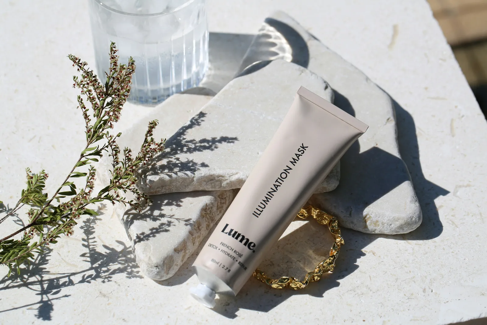 Beige skincare tube resting on stacked stone slabs beside a glass of water, a gold bracelet and a sprig of dried flowers in sunlight