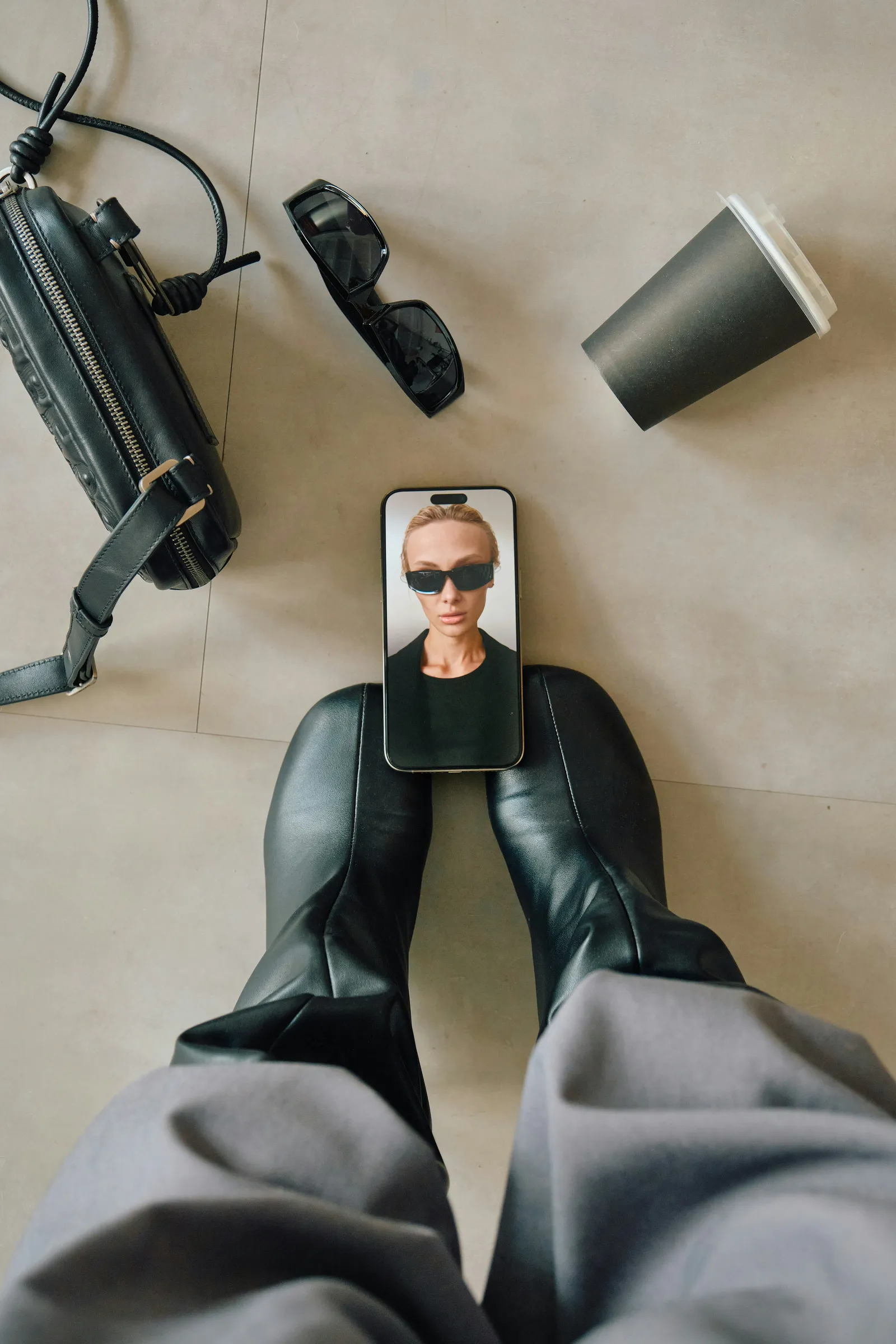 Top-down view of a smartphone showing a selfie resting between leather boots, next to a black handbag, sunglasses and a paper coffee cup