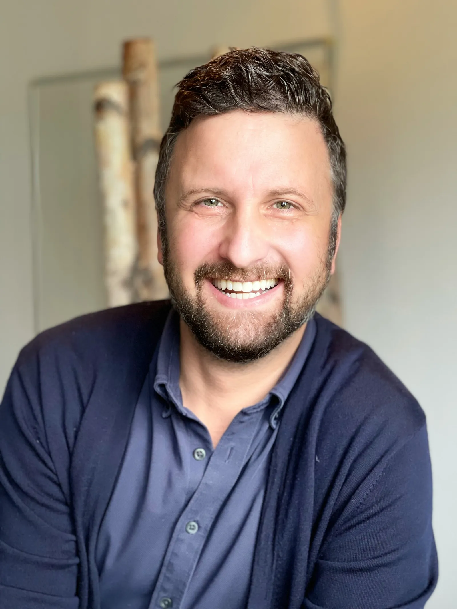 Smiling bearded man in a navy cardigan over a blue shirt, photographed indoors with birch trunks behind him
