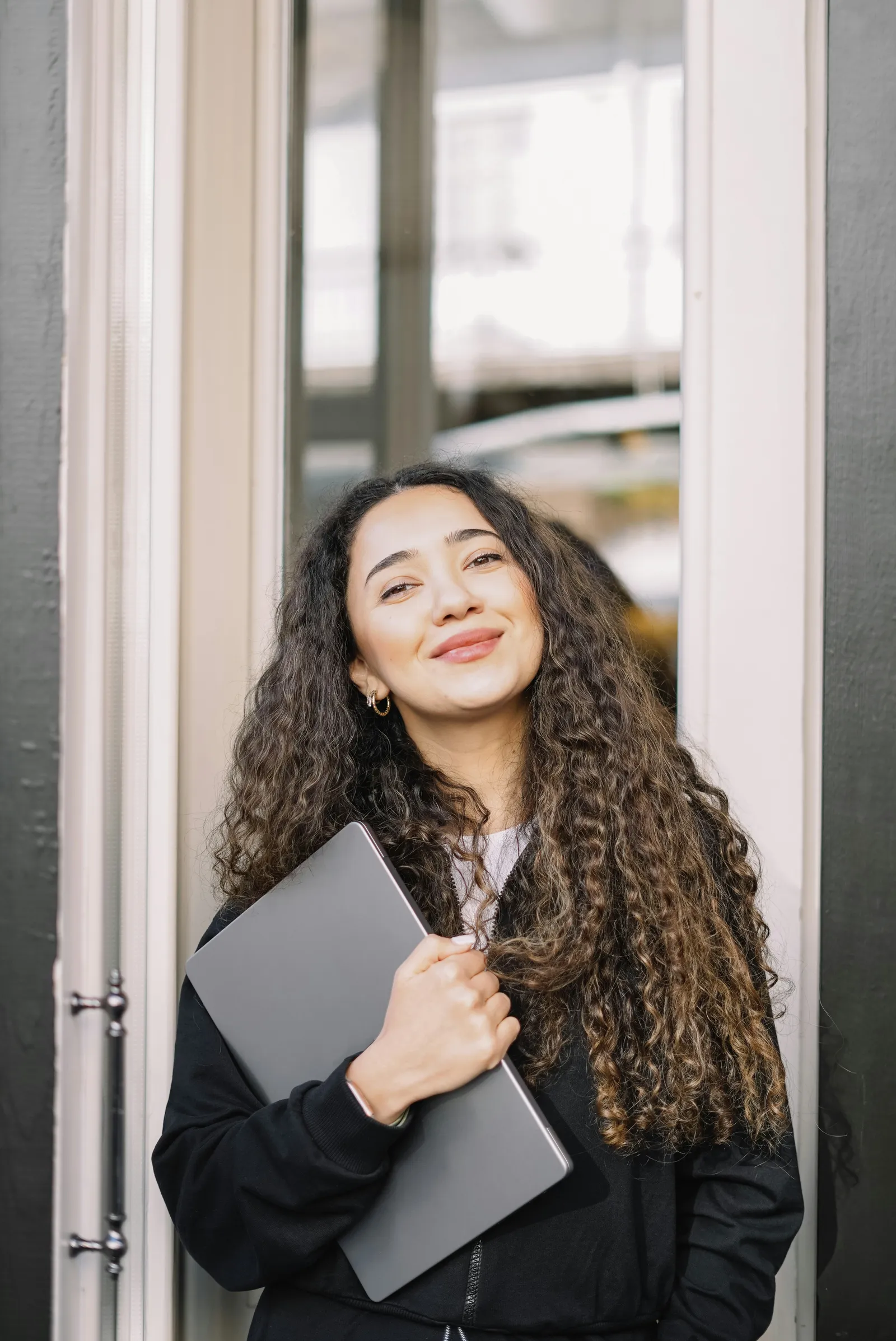 Smiling woman with long curly hair holding a closed laptop in a black jacket in front of a glass door