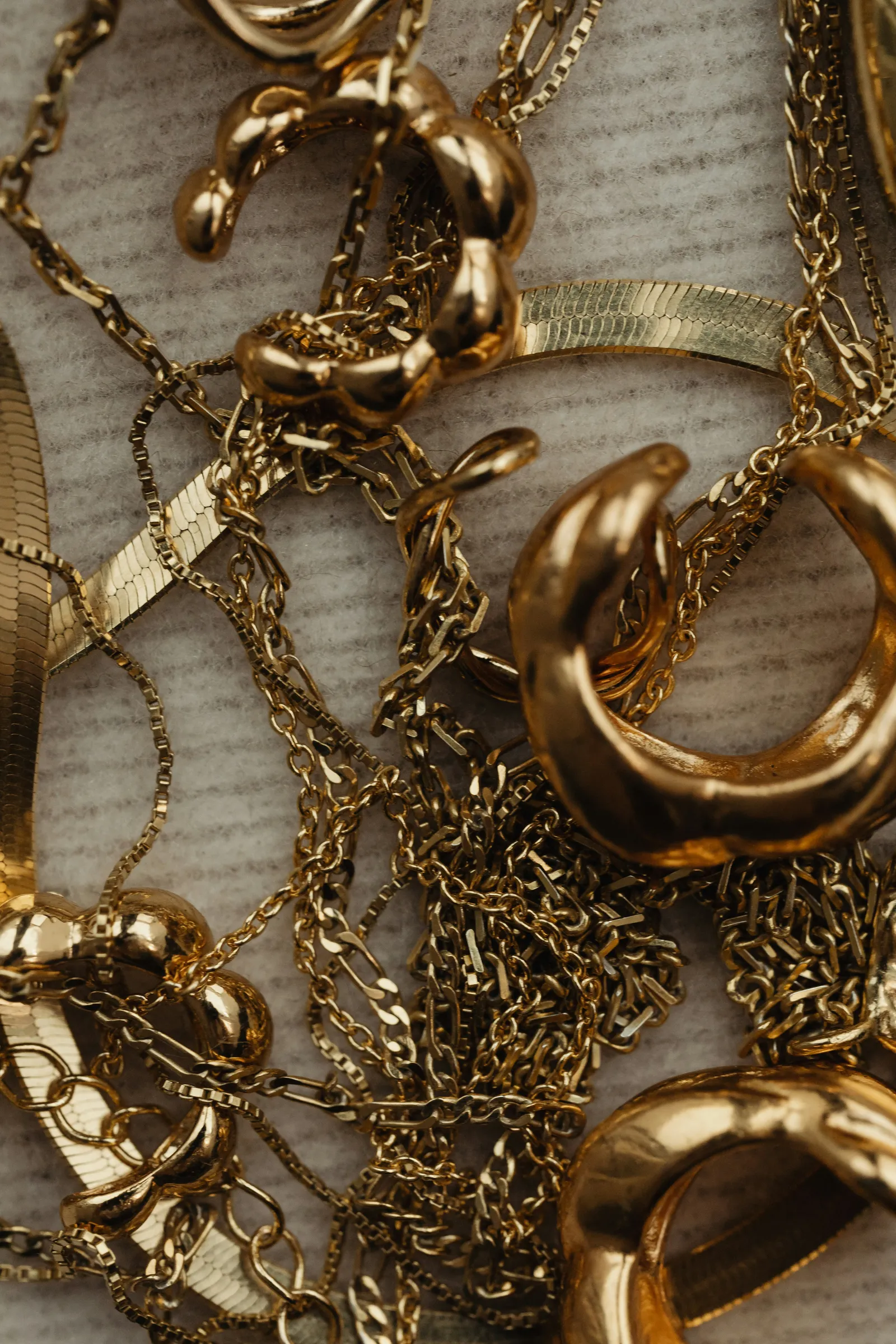 Close-up of tangled gold chains, hoop earrings and rings piled on a textured cream surface
