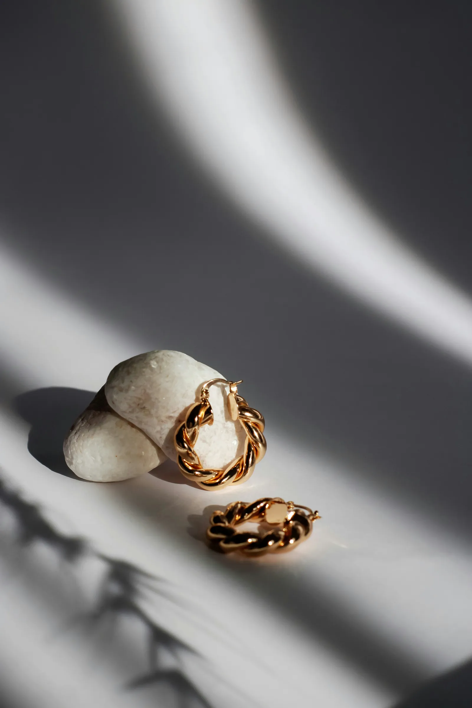Pair of twisted gold hoop earrings resting on white pebbles in dramatic light and shadow