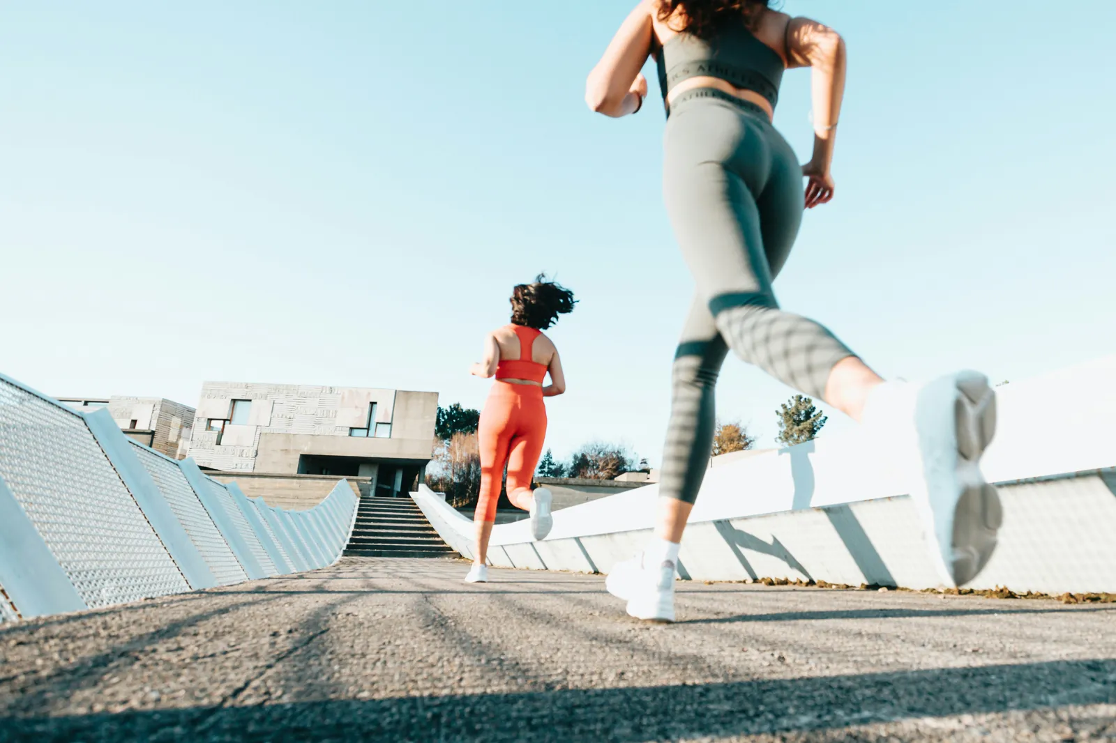 Two women in leggings and sports tops running up a concrete walkway on a sunny day, seen from behind