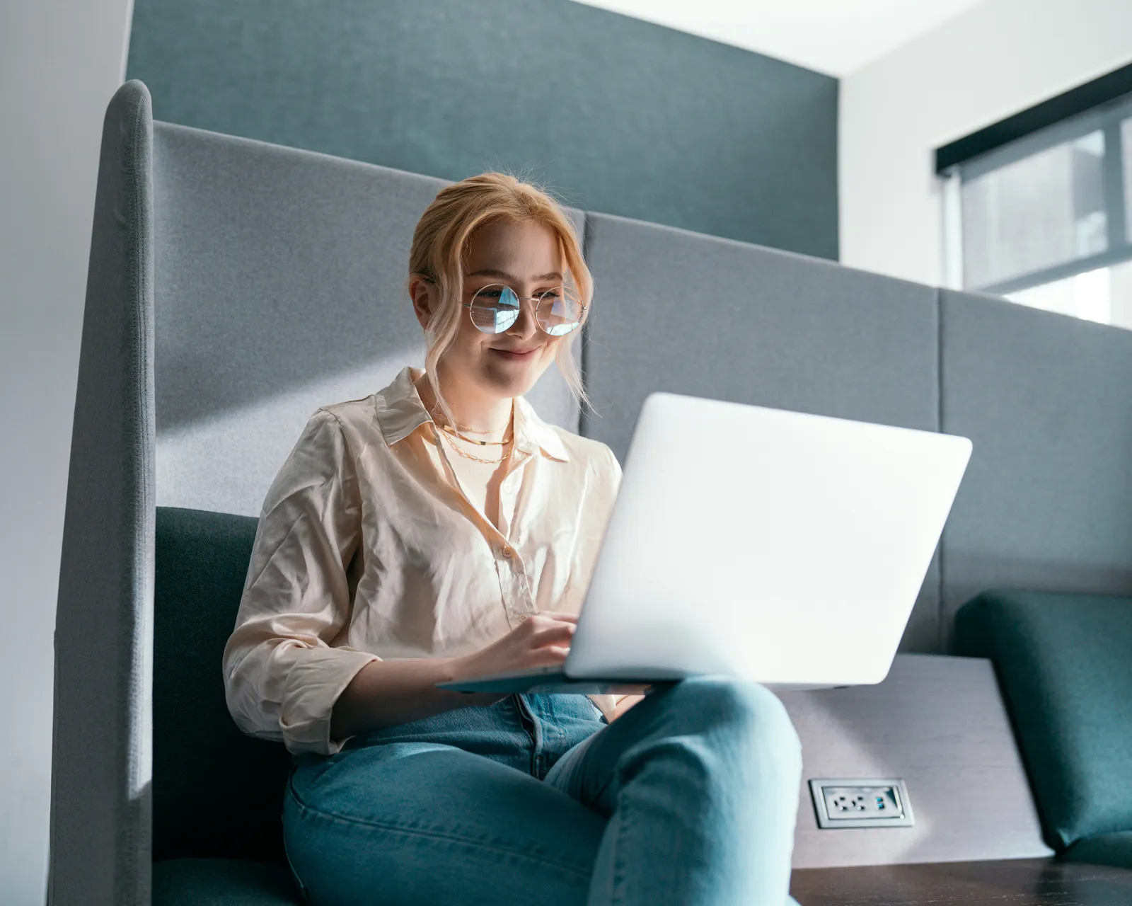 Smiling woman with glasses working on a laptop in a grey upholstered office booth by a window