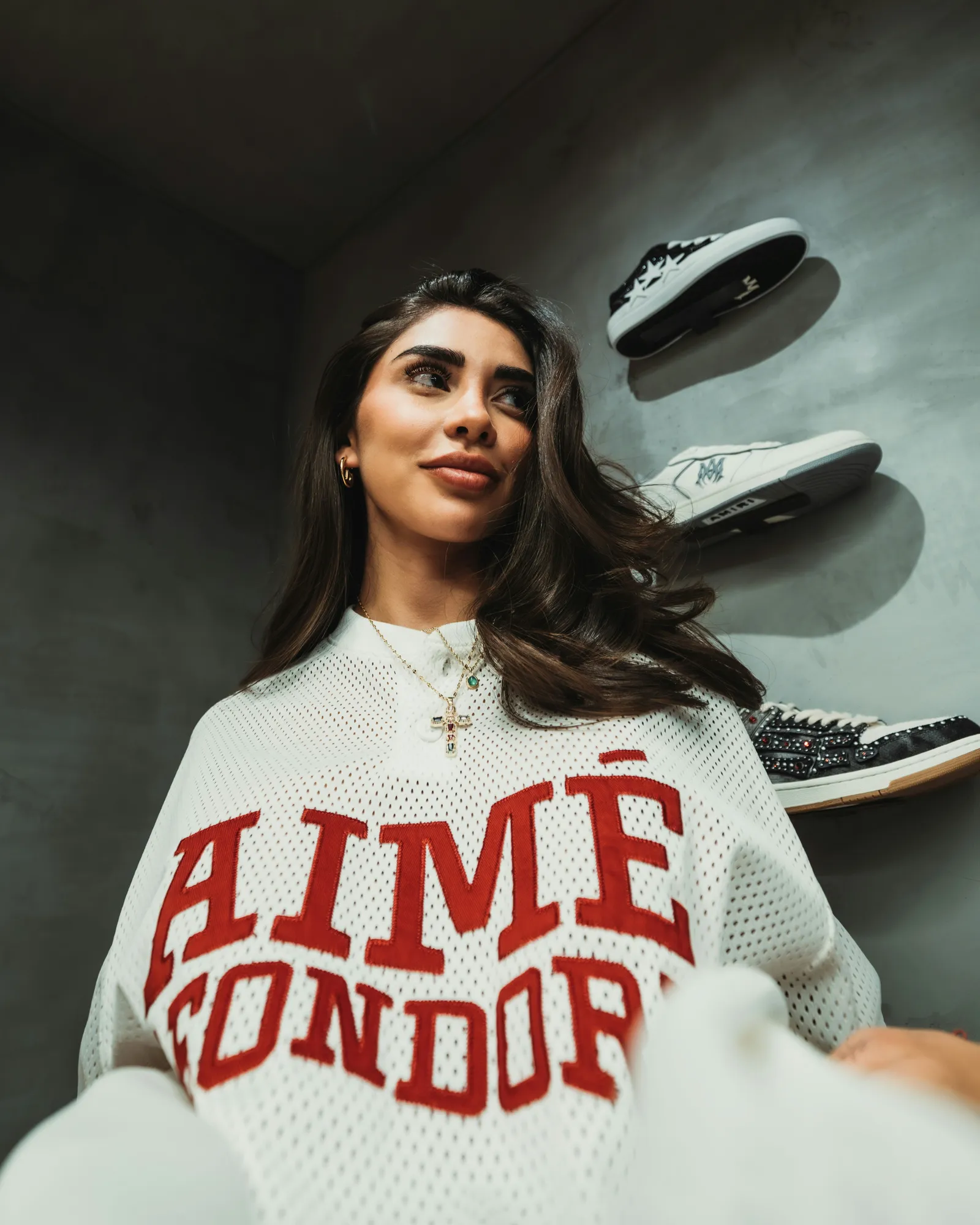 Low-angle photo of a woman in a white mesh jersey with red lettering smiling in a concrete store with sneakers displayed on wall shelves