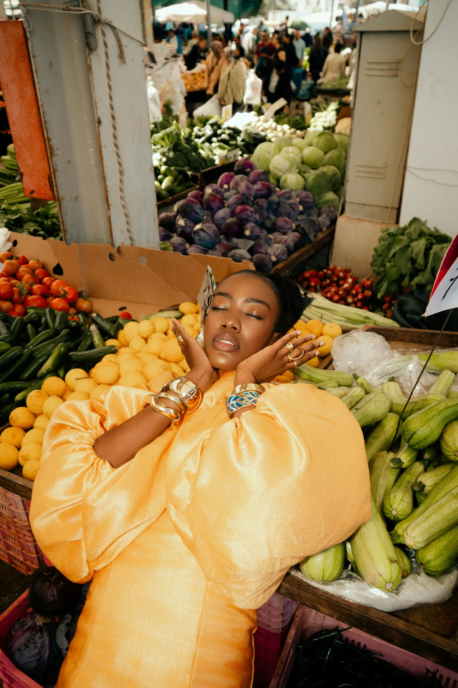 Woman in a shiny orange puff-sleeve dress and gold bangles lying with eyes closed across a vegetable market stall