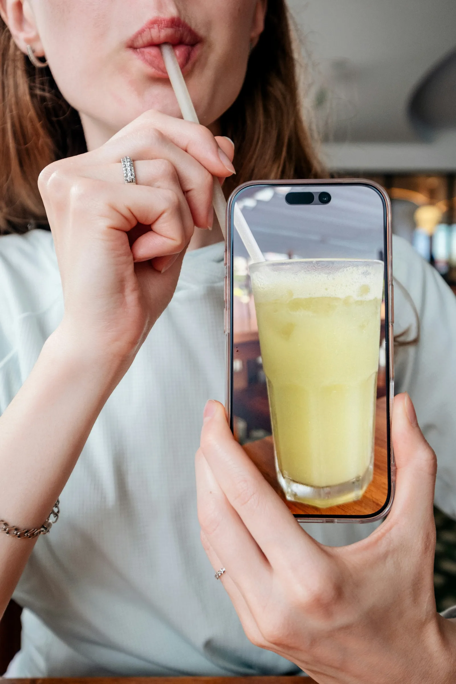 Woman drinking a yellow smoothie through a straw while holding a smartphone that shows the same glass on its screen