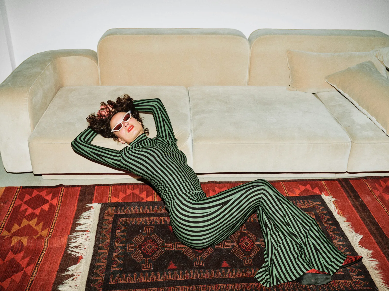 Woman in a green and black striped dress and white sunglasses lying back across a beige velvet sofa onto patterned red rugs